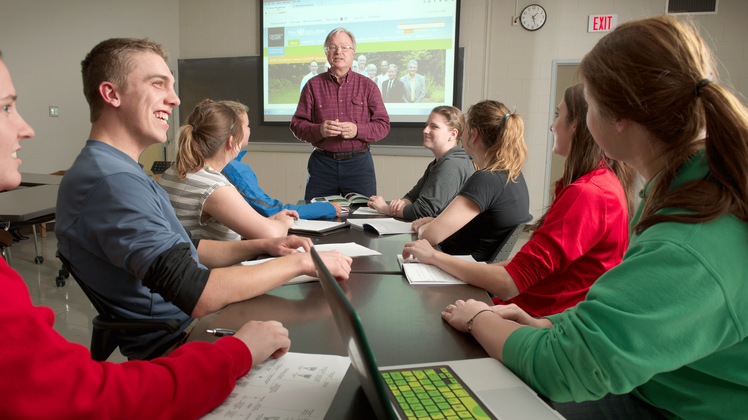 Students sit at a table listening to professor, with a website projected on a screen behind him