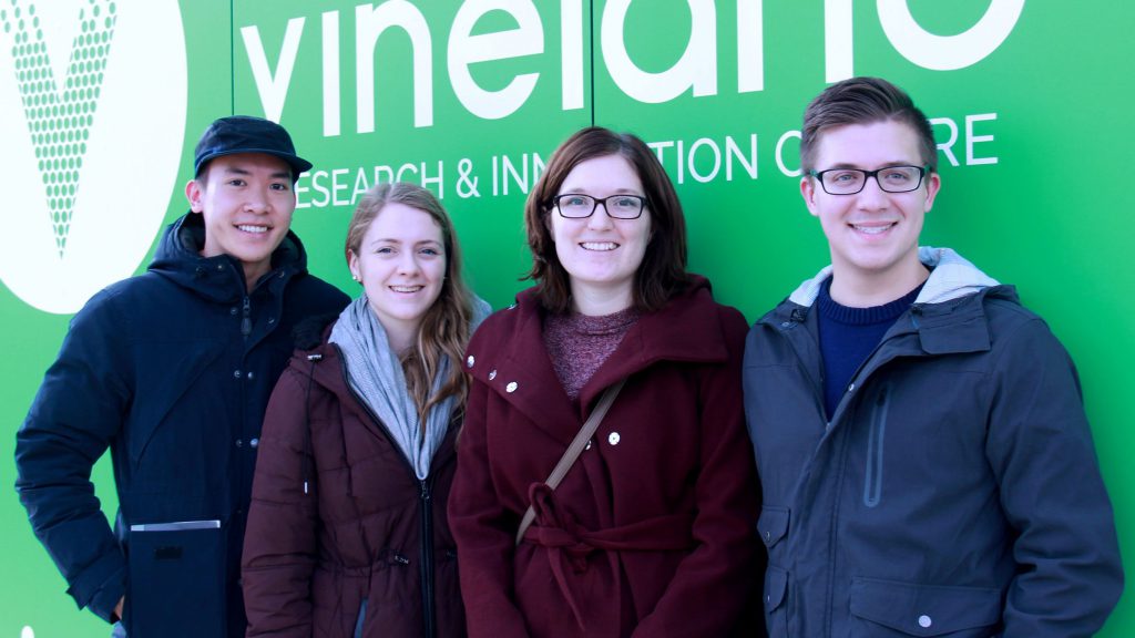 Four students stand in front of green sign at greenhouse