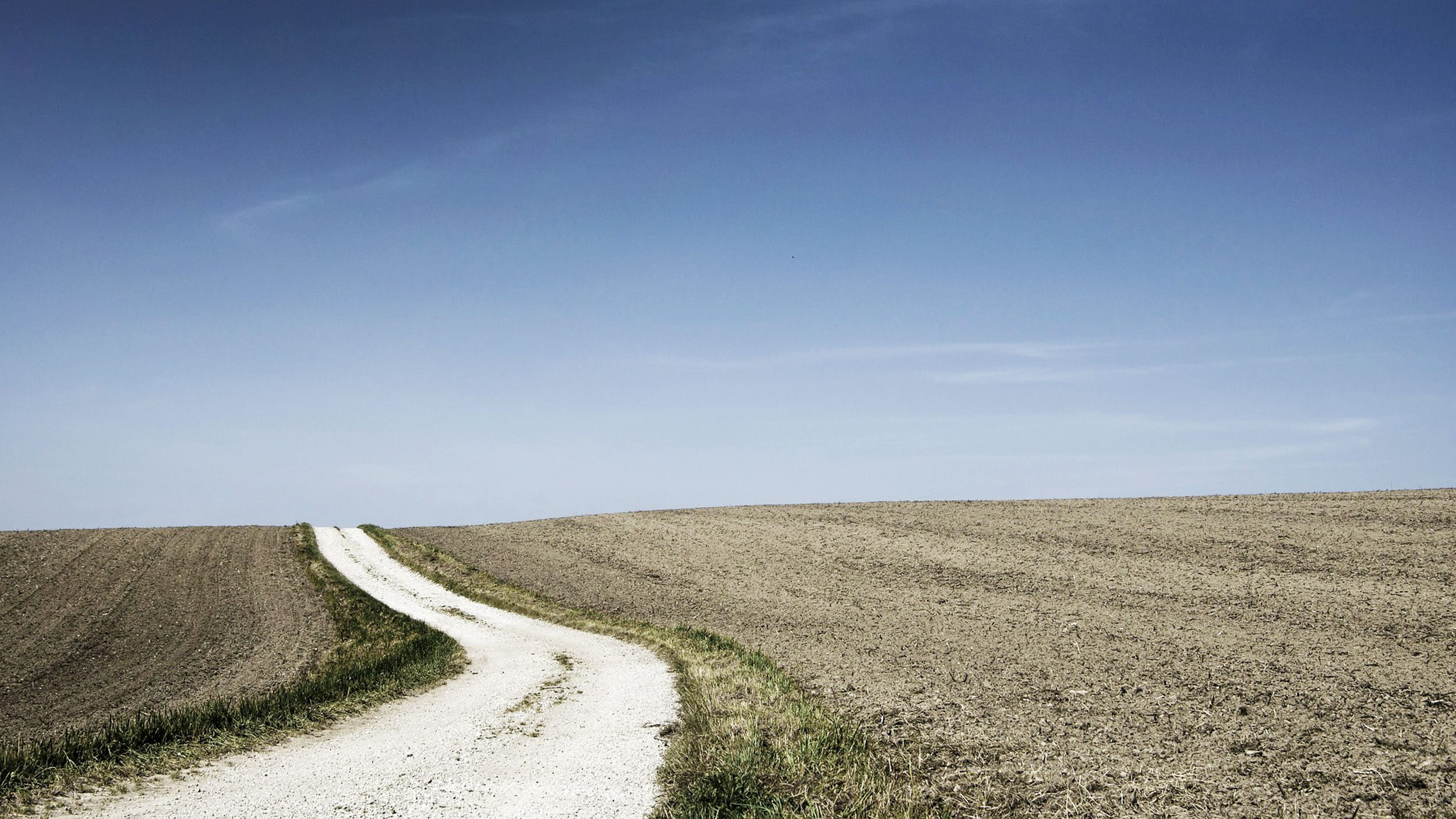 A dirt road runs through a bare field under a blue sky