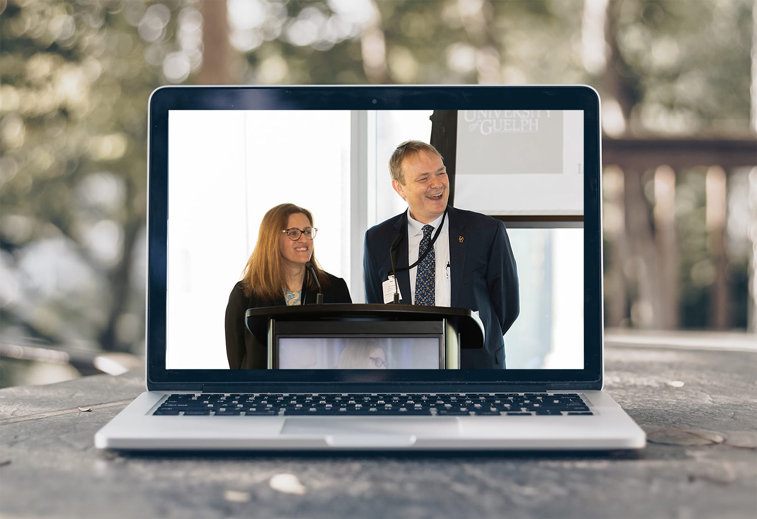 A laptop sits on an outdoor table, with an image of two MCs at a podium