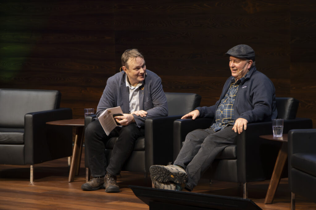 Two men sit on stage in armchairs during a discussion. One holds notes while the other gestures, both smiling. A small table and glasses of water sit between them.