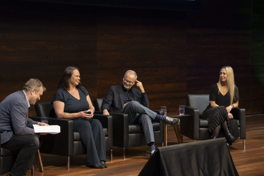 Four panelists sit on stage in black armchairs during a discussion. Three are facing the audience, one is looking down at notes. The stage has a dark wood backdrop and small glasses of water on side tables.