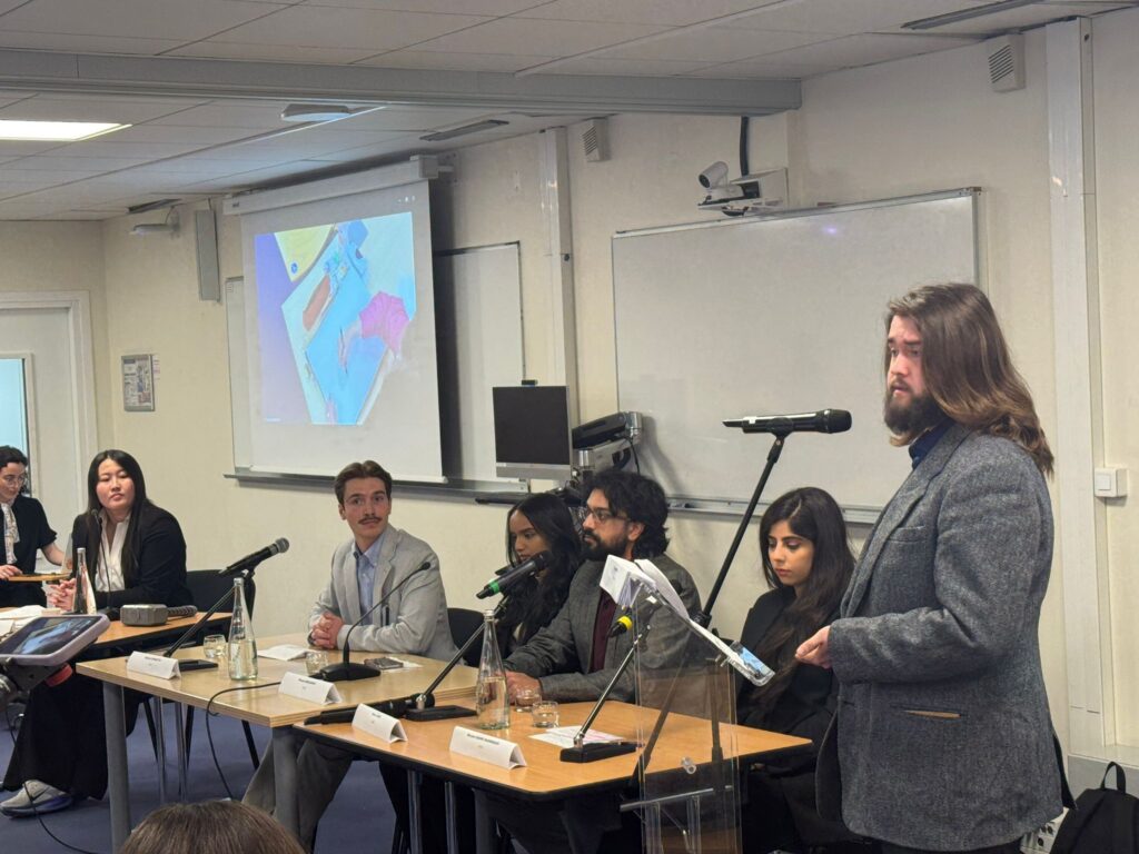 Tristan Cook speaks into a microphone in front of a seated panel of peers